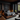 A wide shot of a luxurious, modern bedroom with a dark and moody color scheme. A large platform bed with charcoal grey bedding rests centrally, flanked by dark wood paneling and two warm wall sconces. To the right, expansive glass doors open to an outdoor deck with a fire pit and a lush forest of tall evergreen trees, while a live-edge wood table and armchair are in the foreground.