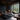 A luxurious and modern bedroom with dark charcoal and black decor. A large, low-profile bed with soft grey bedding sits to the left on a dark wooden platform. A contemporary glass fireplace with a burning fire and neatly stacked wood is visible in the middle. Expansive floor-to-ceiling windows offer a stunning view of a dense green pine forest, with a dark grey chaise lounge positioned to face the view. A rustic, textured wooden table with dark coffee mugs and books is in the foreground on the right.