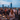 A group of people are seen dining outdoors on a city rooftop at dusk. In the foreground, a man and a woman are smiling and laughing across a wooden table filled with food, drinks, and lit candles, with string lights strung above them. In the blurred background, other diners and the distant lights of a city skyline are visible under a deep blue sky.
