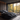 A luxurious modern bedroom features a large, dark gray upholstered bed with multiple pillows and a blanket, positioned against a dark brown vertical wood-paneled wall. Floor-to-ceiling windows dominate the wall opposite the bed, offering a stunning view of an autumnal forest filled with vibrant yellow and orange trees. Outside on a small balcony, a dark armchair and a round table with mugs sit, providing a cozy spot to enjoy the natural scenery.