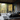 A luxurious modern bedroom interior with a large bed featuring dark grey bedding and a fluffy grey rug, facing expansive floor-to-ceiling glass windows. Beyond the windows, an outdoor balcony overlooks a vibrant autumn forest with trees displaying bright yellow, orange, and green foliage. The room's interior includes dark wood plank walls and ceiling, a minimalist dark wood bedside table with a glowing lamp, and potted plants near the window, creating a serene and elegant retreat.