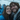 A close-up portrait of a young man and woman smiling happily and looking directly at the camera, sharing a clear umbrella covered in raindrops. The woman on the left wears a bright blue rain jacket and has long wet brown hair. The man on the right has a beard, wet brown hair, and wears a grey hoodie under a dark rain jacket. Blurred figures in similar blue jackets are visible in the background, suggesting an outdoor event on a rainy day.