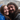 A close-up portrait of a happy young couple embracing under a clear umbrella during a rainy day. The woman on the left wears a bright blue rain jacket and smiles broadly, while the man on the right, with long dark hair and a beard, also smiles, with rain droplets visible on their hair and faces. The background is softly blurred, showing hints of other people and umbrellas.