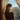 A full-length vertical shot from behind shows a person, likely a woman, with long, wavy blonde hair standing by a window. She is silhouetted against a bright, sun-drenched sky, with warm golden light creating a beautiful rim light on her hair and shoulders, and a sun flare obscuring part of her face. Beyond the sheer, patterned curtains framing the window, a clear blue sky, distant mountains, and an urban landscape of houses are visible.