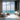 A luxurious modern bathroom features floor-to-ceiling windows overlooking a vast turquoise ocean and a distant white-roofed coastal village. Two contemporary white freestanding bathtubs sit on a glossy white marble floor, bathed in natural light. To the right, a dark textured wall supports a floating marble vanity with a vessel sink, decorative candles, and four glass pendant lights, while the left side includes vertical wooden wall panels and a rustic wooden stool with a diffuser.