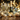 Two clear champagne flutes filled with sparkling golden champagne are prominently displayed on a dark wooden table. In the soft, blurred background, a green champagne bottle with a gold label is visible, surrounded by a scattering of golden and silver Christmas ornaments, decorative tinsel, and fine glitter, all set against a dark backdrop illuminated by warm, glowing golden bokeh lights. The scene conveys a luxurious and festive celebratory atmosphere.