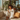 A smiling woman with curly hair sits cross-legged on a beige couch, bathed in sunlight. She wears white pajamas with gold trim and holds a delicate teacup. Behind her, a large window reveals a cityscape and an abundance of indoor plants, creating a warm and inviting atmosphere.