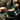 A close-up, warm-toned image features a dark teal ceramic mug filled with frothy coffee on a matching saucer, accompanied by a gold spoon. To the left, a stack of golden-brown cookies rests on a small silver plate. In the blurred background, lit candles in green holders, pine branches, and pine cones create a cozy, festive atmosphere, with scattered gold glitter on a dark, mottled surface.