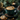 A close-up, slightly angled shot of a latte with detailed leaf art in a dark teal ceramic mug, resting on a matching saucer filled with scattered coffee beans. To the left, two chocolate chip cookies and a small golden spoon are arranged on a dark grey fabric. The background features blurred pine branches, pinecones, golden holiday ornaments, and a soft, glowing candle, creating a warm and festive atmosphere.