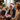 A cheerful elderly man with a white beard and a red Santa hat laughs joyfully, his mouth wide open, while holding a patterned Christmas gift box tied with a bright red ribbon. He is seated on a patterned rug, surrounded by blurred family members, including children, and a softly lit Christmas tree with golden lights in the background, creating a warm and festive home scene.