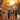 Five young girls in Halloween costumes walk away from the viewer down a residential street covered in autumn leaves. The girls, dressed as various kinds of witches, carry pumpkin-shaped candy buckets. The scene is bathed in warm, golden light from the setting sun, and decorated with lit streetlights and jack-o-lanterns on the curbside.