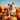 A happy bulldog puppy sits proudly atop a mound of bright orange pumpkins. The dog is white and tan, wearing a patterned collar, and is looking directly at the viewer with its tongue slightly sticking out, giving it a playful expression. The pumpkins are various sizes and shapes, creating a dynamic composition against a clear blue sky.