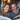 A mature couple, a blonde woman and a gray-haired man, sit side-by-side, both looking down and laughing. The woman wears a brown sweater, while the man is in a gray sweater over a striped shirt. In the blurred background are two round, glowing lamps that suggest a warm, intimate setting.