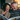A middle-aged woman and man are sitting closely together, engrossed in a magazine. The woman, wearing a knitted sweater, is laughing heartily with her eyes closed. The man, with graying hair, is smiling broadly, his eyes sparkling with amusement as he looks at the magazine. The warm, indoor setting creates a sense of intimacy and joy.