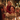 A blonde little girl in a red knitted sweater sits on a man's shoulders, both laughing joyfully at a festive, candlelit dinner table. The man, with dark hair and a patterned beige sweater, smiles broadly, surrounded by other family members blurred in the foreground and background. Warm, glowing Christmas lights create a cozy and celebratory atmosphere, highlighting the genuine connection and happiness shared during the holiday meal.