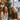 A medium shot captures two young women, one with red hair and another with blonde curly hair, laughing heartily at a wooden table in a festive cafe. In front of them are a latte, a slice of cake, and glowing candles, with a decorated Christmas tree and string lights in the warm, blurred background, and a street scene visible through a window. The image conveys genuine joy and a cozy holiday atmosphere.