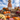 A medium shot displays a whimsical scarecrow standing on stacked hay bales. The scarecrow is wearing an orange plaid shirt, white pants, and brown boots, and has a smiling face painted on its pumpkin head. Pumpkins and potted mums of various colors surround the base of the hay bales creating a festive fall scene with a clear blue sky and soft white clouds in the background.