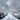 A vertical, eye-level photograph looking down a snow-covered residential street under an overcast sky. Rows of parked cars, completely blanketed in thick white snow, line both sides of the street, which shows a partially cleared, wet, dark grey pavement down the middle. Bare tree branches, also heavily covered in snow, arch overhead, creating a natural tunnel-like effect and framing the peaceful, wintery scene with utility poles and wires visible on the left.