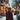 A vertical shot from behind a woman with light brown hair styled in a bun, wearing a dark red, possibly velvet, winter coat. She is walking forward through a snowy environment. Numerous out-of-focus warm golden and orange string lights illuminate the blurry urban background, creating a festive bokeh effect, while white snowflakes fall gently around her. The ground is covered in snow, suggesting a serene winter evening in a decorated city street or market.