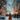 A young girl with long, wavy blonde hair and a vibrant burnt orange winter coat and beanie walks away from the viewer down a snow-covered street. The scene is illuminated by numerous soft, glowing orange and yellow string lights decorating bare tree branches and distant buildings, with countless blurred snowflakes falling, creating a magical, dreamlike winter atmosphere.