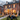 The exterior of a red brick house is decorated extensively for Halloween. A large black spider sits on the front lawn, with various skeletons, pumpkins, and other Halloween decorations arranged around the yard and on the front porch. The house's windows are illuminated from the inside, creating a warm and inviting, yet slightly spooky, ambiance.
