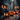 A Halloween scene shows a dark, weathered wooden table with a black, vintage-style lantern on the left holding two lit candles. Next to the lantern are three orange pumpkins with carved, illuminated faces. In the blurred background, there is a building and bokeh lights, creating a spooky and festive atmosphere.