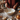 A close-up shot of a person wearing a rust-colored knit sweater, with their hands holding a whisk in a large cream-colored bowl containing white flour and a yellow liquid. The scene is set on a wooden table, decorated with small white pumpkins, dried brown leaves, and a cream lace placemat, evoking a warm and autumnal baking atmosphere.