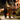 A photograph shows three young children in Halloween costumes walking side-by-side on a concrete sidewalk at night. From left, one child wears a black skeleton costume and holds a flashlight, the middle child wears a full orange and black tiger suit and holds an illuminated jack-o'-lantern bucket, and the child on the right wears a black and orange leopard print top with a tutu skirt and striped tights. In the blurred background, houses are adorned with warm, glowing Halloween decorations and pumpkins.