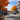 A wide-angle view of a charming small town street during autumn, lined with historic buildings featuring storefronts and vibrant orange and gold maple trees. Cars are parked along both sides of the asphalt street, which shows shadows from the trees and buildings. The sky above is a clear blue with scattered white clouds, creating a bright and peaceful fall day atmosphere in a classic American village.