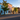 A picturesque view of a small town street featuring a row of charming, vibrantly colored historic buildings under a clear blue sky. The scene is illuminated by the soft, golden light of morning or late afternoon, casting long shadows across the empty asphalt road and sidewalk. Prominent buildings include a bright blue one, a salmon-pink one, and a golden-yellow one, all adorned with trees and flower baskets, evoking a peaceful and inviting atmosphere.
