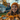 A young woman with a wide, joyful smile, wearing an orange knit beanie and matching sweater, looks upward and to the left. She is in the foreground, with a pile of vibrant golden and brown autumn leaves on a grey surface directly in front of her. In the soft-focus background, a bearded man is visible, holding a long-handled broom or rake, standing near a white house with trees displaying beautiful fall foliage under a soft blue sky.