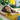 A young woman with long, wavy brown hair lies peacefully in a light-colored woven hammock, with her eyes closed and a gentle smile. She is wearing a yellow t-shirt and blue jeans, illuminated by warm sunlight. The background is a softly blurred outdoor garden featuring lush green foliage and a bright yellow palm frond, creating a serene summer atmosphere.