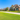A wide-angle, eye-level view of a tranquil suburban street lined with multiple modern, two-story homes under a clear bright blue sky. The foreground features a vast, vibrant green lawn and a light-colored concrete sidewalk that curves gently, leading towards the detailed facades of the houses. Each home showcases a blend of brick, stone, and stucco, with gabled roofs, well-maintained shrubbery, and trees casting subtle shadows on the impeccably kept lawns, conveying an atmosphere of peace and prosperity.