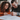 A young adult couple, a woman with curly brown hair and a man with a beard, both wearing dark-rimmed glasses, are seated at a warm wooden table. They are both smiling brightly as they hold pens and sign a white document. Natural light illuminates the scene, highlighting their engaged expressions and a cozy indoor setting with a dark green wall and window in the background.