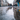 A low-angle photograph captures a bustling city street on a grey, rainy day. The foreground features a large, glistening puddle that perfectly reflects the upper half of the scene, showing blurred ornate buildings, tall streetlights, and bare trees. Numerous pedestrians in winter coats walk away from the camera on the wet sidewalk, their legs and reflections prominently visible, conveying a sense of continuous urban movement.