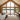 A wide shot of a modern, light-wood paneled cabin living room with a vaulted ceiling and dark gray flooring. The room features expansive floor-to-ceiling glass doors and triangular upper windows that offer a panoramic view of a calm lake surrounded by dense green forest. A large light beige sectional sofa sits on the left, a natural fiber rug covers the center, and a wooden armchair with a dark blanket is on the right, all bathed in soft natural light.