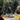 Three diverse women are joyfully gathered around a rustic wooden table outdoors, engaged in lively conversation and sharing a meal. The woman in the center, with dreadlocks and a denim jacket, is captured mid-laugh, while another woman with curly blonde hair smiles warmly at her. Various dishes, including a fresh salad, and amber-colored drinks are visible on the table, set against a lush green garden backdrop under natural daylight.