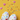 A vibrant overhead flat lay photograph showcases two white eggs decorated with numerous small, multi-colored heart shapes on a bright yellow surface. Three pink puffy hearts and scattered heart-shaped confetti in various colors float above and around the eggs, while fresh white daisy-like flowers with yellow centers are visible in the bottom left corner, creating a joyful and celebratory scene.