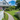 A picturesque gray shingled house with a white front porch features an American flag hanging by the entrance. A classic white picket fence lines the foreground on the left, leading to a vibrant, colorful summer garden. A winding gravel path cuts through a bright green lawn, leading towards the front door under a clear blue sky with fluffy white clouds.