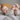 A cheerful baby, approximately six to twelve months old, is lying on their back on a light grey textured bedspread. The baby has fair skin, bright blue eyes, and a wide smile, looking directly at the viewer while playfully holding their bare feet with both hands. In the soft-focus background, a warm orange cushion and a lighter pillow are visible, contributing to the cozy and inviting bedroom setting.