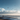 A wide outdoor shot depicting a man in silhouette walking barefoot on a wet sandy beach towards the ocean on the left side of the frame. The sky above transitions from bright blue at the top to fluffy white and grey clouds in the middle, and a brilliant, hazy golden glow near the horizon. A large flock of birds, also silhouetted, flies dynamically across the mid-right sky, adding movement to the vast, peaceful coastal scene.