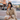A stylish East Asian woman with a bob haircut and bangs, wearing a beige trench coat, sits on a wooden bench outdoors. She is holding a piece of street food, resembling a taco or savory pastry, near her mouth, with a take-out box resting on her lap, against a blurred backdrop of historic brick buildings and a bustling street scene with distant mountains under a cloudy sky. The woman looks directly at the viewer with bold red lipstick, embodying a casual yet fashionable urban traveler enjoying local cuisine.