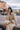 A stylish East Asian woman with a bob haircut and bangs, wearing a beige trench coat, sits on a wooden bench outdoors. She is holding a piece of street food, resembling a taco or savory pastry, near her mouth, with a take-out box resting on her lap, against a blurred backdrop of historic brick buildings and a bustling street scene with distant mountains under a cloudy sky. The woman looks directly at the viewer with bold red lipstick, embodying a casual yet fashionable urban traveler enjoying local cuisine.