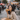 A stylish Asian woman with long dark hair, wearing a beige trench coat and red lipstick, sits on a weathered wooden bench. She holds a hard-shell taco to her mouth, looking directly at the viewer, while a brown cardboard box with more tacos rests on her lap. The background is a soft blur of a busy urban street or outdoor market with people, buildings, and market stalls.