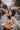 A stylish Asian woman with long dark hair, wearing a beige trench coat and red lipstick, sits on a weathered wooden bench. She holds a hard-shell taco to her mouth, looking directly at the viewer, while a brown cardboard box with more tacos rests on her lap. The background is a soft blur of a busy urban street or outdoor market with people, buildings, and market stalls.