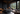A luxurious and modern bedroom with dark charcoal and black decor. A large, low-profile bed with soft grey bedding sits to the left on a dark wooden platform. A contemporary glass fireplace with a burning fire and neatly stacked wood is visible in the middle. Expansive floor-to-ceiling windows offer a stunning view of a dense green pine forest, with a dark grey chaise lounge positioned to face the view. A rustic, textured wooden table with dark coffee mugs and books is in the foreground on the right.