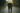 A couple is depicted from behind, walking away from the viewer on a wet, paved country road during a gentle rain. They share a bright yellow umbrella, and the man has his arm around the woman's shoulder. The scene conveys intimacy and togetherness, with lush, muted green and brown trees lining the road under an overcast sky.