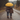 A couple, viewed from behind, walks down a wet, unpaved road in a lush forest during a rain shower. They are holding a bright yellow umbrella, and the man has his arm around the woman's shoulder as they walk together, creating an intimate scene. Both are wearing plaid flannel shirts and boots, with trees showing muted autumn colors in the soft, diffused light of the background.
