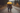 A couple, viewed from behind, walks down a wet, unpaved road in a lush forest during a rain shower. They are holding a bright yellow umbrella, and the man has his arm around the woman's shoulder as they walk together, creating an intimate scene. Both are wearing plaid flannel shirts and boots, with trees showing muted autumn colors in the soft, diffused light of the background.