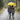 A couple walks away from the viewer on a wet gravel path during an autumn rain shower, sharing a bright yellow umbrella. The man, in a blue and red plaid shirt and jeans, has his arm around the woman, who wears a purple and black plaid jacket and dark pants; both are wearing boots. The scene is set amidst vibrant green and yellow autumn trees, with visible raindrops falling, creating a cozy and intimate atmosphere.
